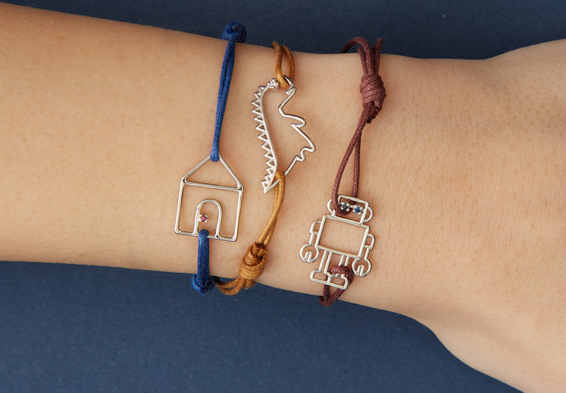 Cord bracelets with small white gold pendants shaped like a house, dinosaur, robot with precious stones on model's wrist