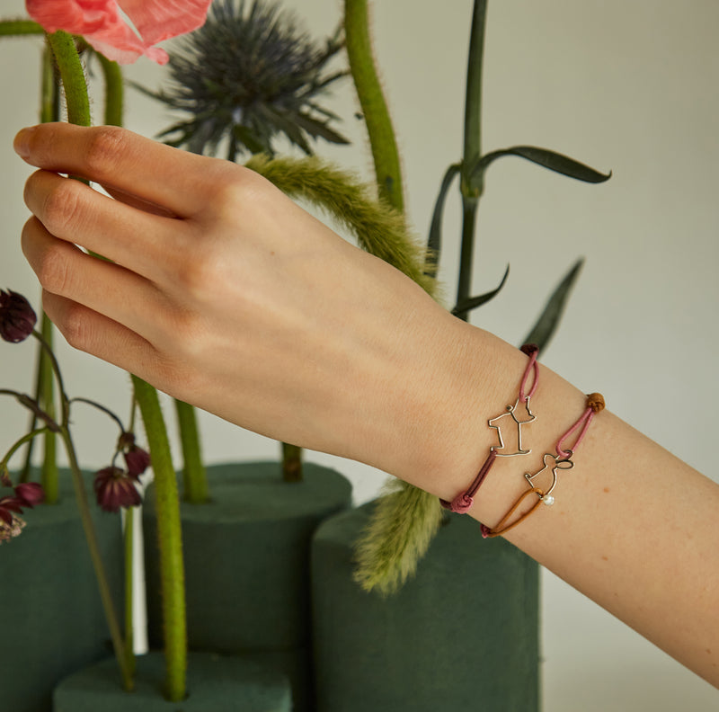 Hand touching flowers wearing two pink cord bracelets with small white gold pendants shaped like a little dog and rabbit