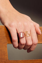 Load image into Gallery viewer, Hand wearing a rabbit shaped gold ring and a gold ring with an eggplant cameo on porcelain
